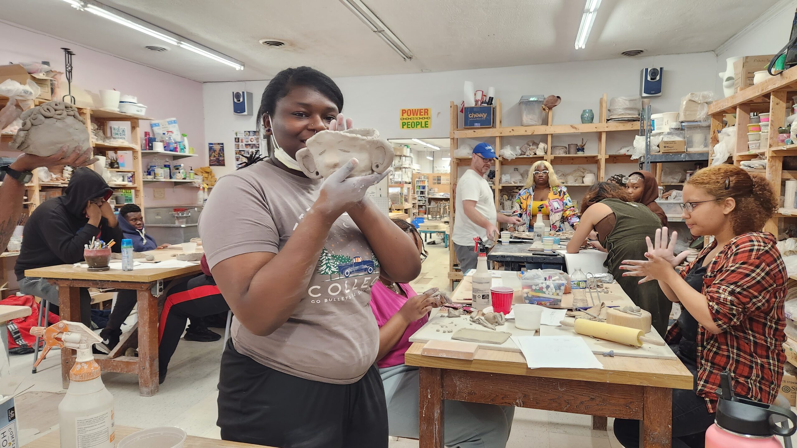 A young artist stand in a ceramics studio holding a work in progress. Other students are working at a long table behind her.