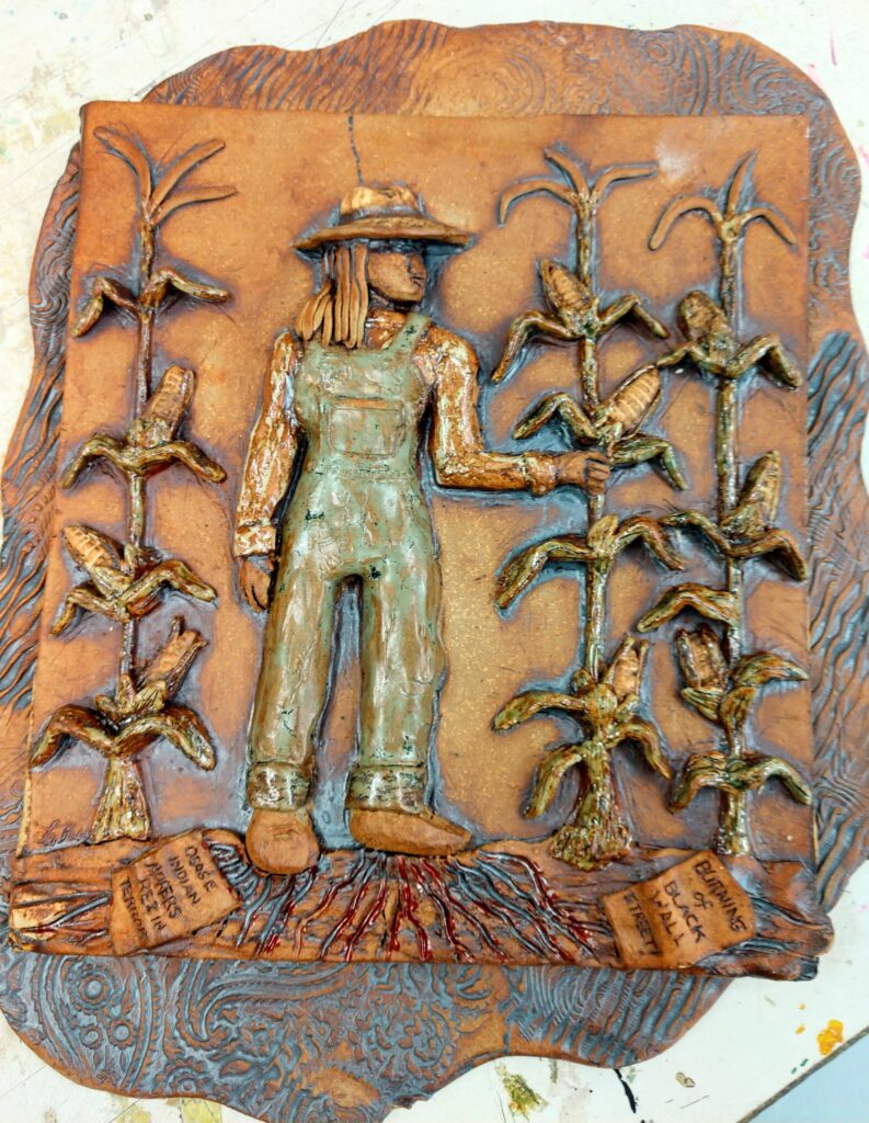 A green and tan ceramic tile showing a figure wearing overalls and a hat standing amid corn stalks.
