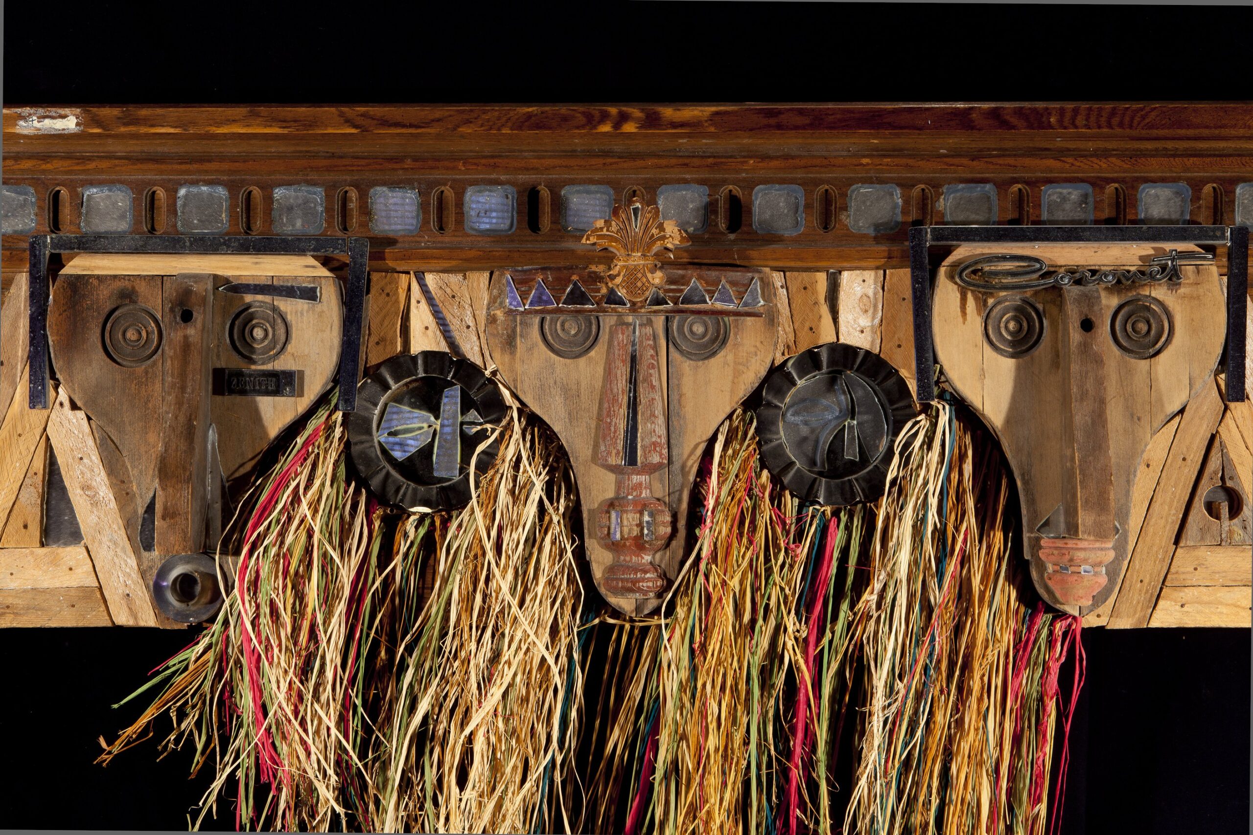 A wood wall sculpture of three faces with multi-colored rafia hanging from between the faces.