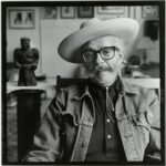 Seated man in hat and jean jacket with glasses and large mustache.