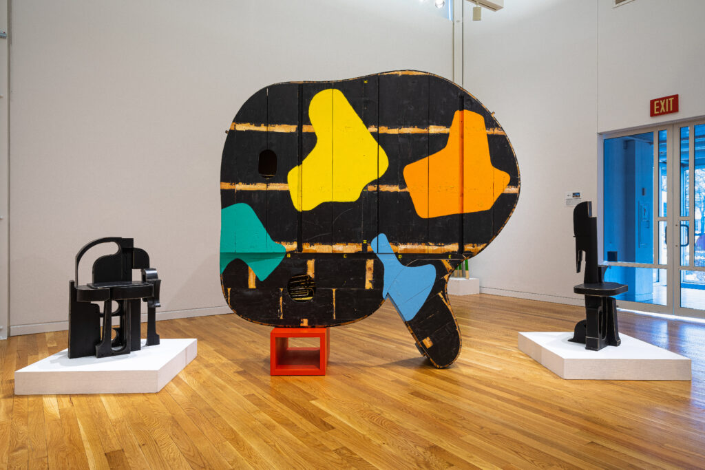 A gallery installation featuring a large wooden sculpture painted black shaped like a grand piano propped up on its side on a small red box. Two smaller sculptures are placed on either side.