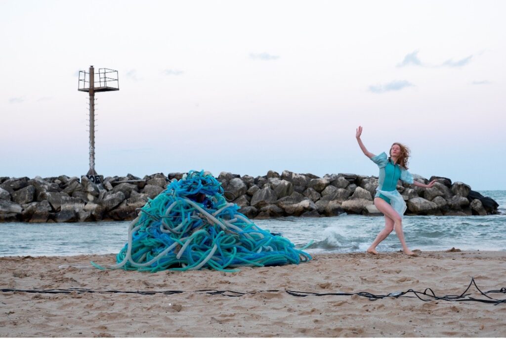 A performer in a blue garment next to an art piece at the Water Music on the Beach event.