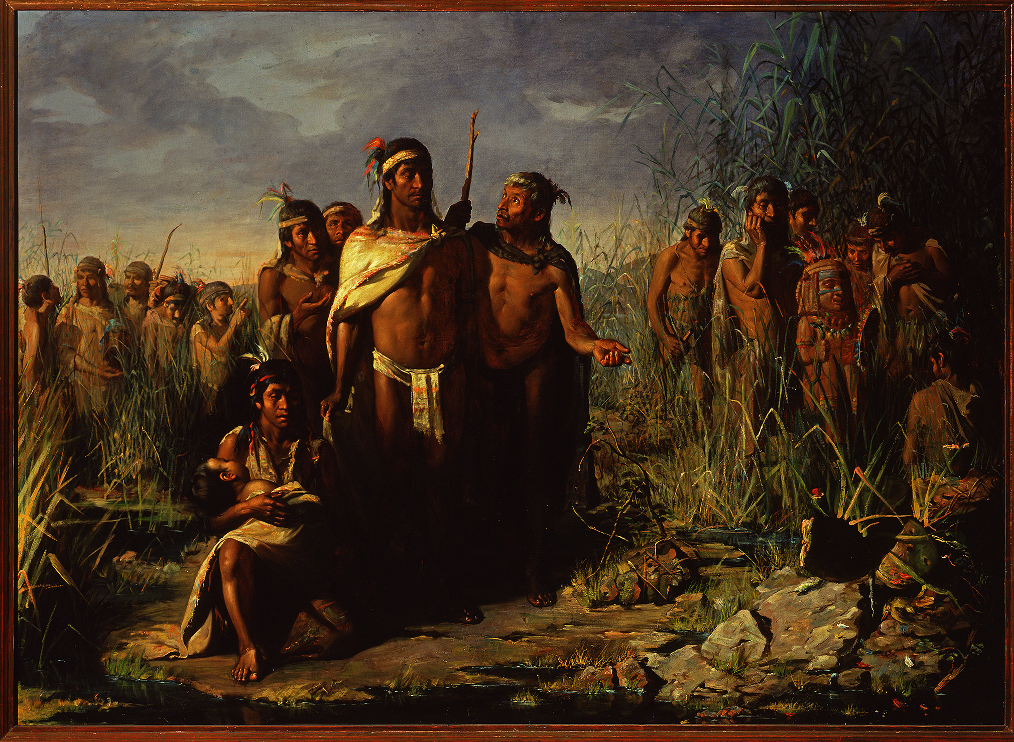 Indigenous people wearing loincloths, headbands, and feathers are assembled outdoors in an area with tall grass, corn, and rocks.