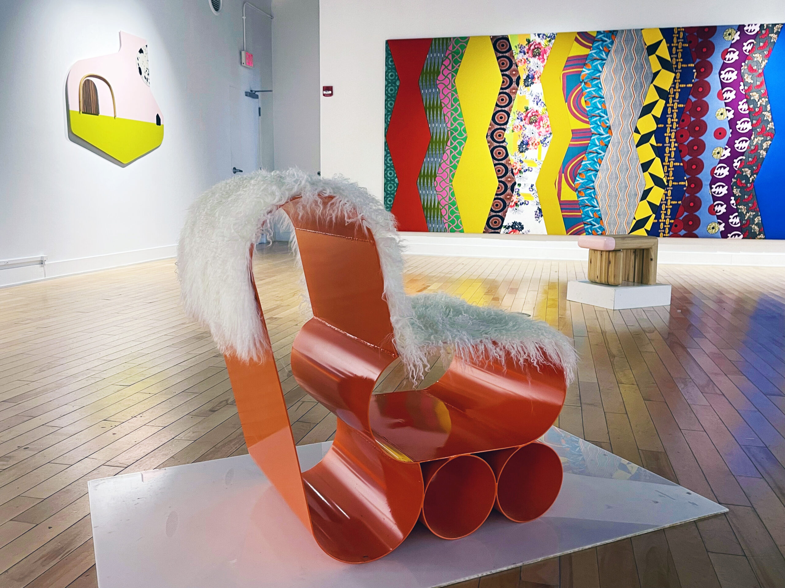 A gallery installation featuring an orange curved metal chair with a white fur covering in the foreground and a brightly colored abstract painting in the background.