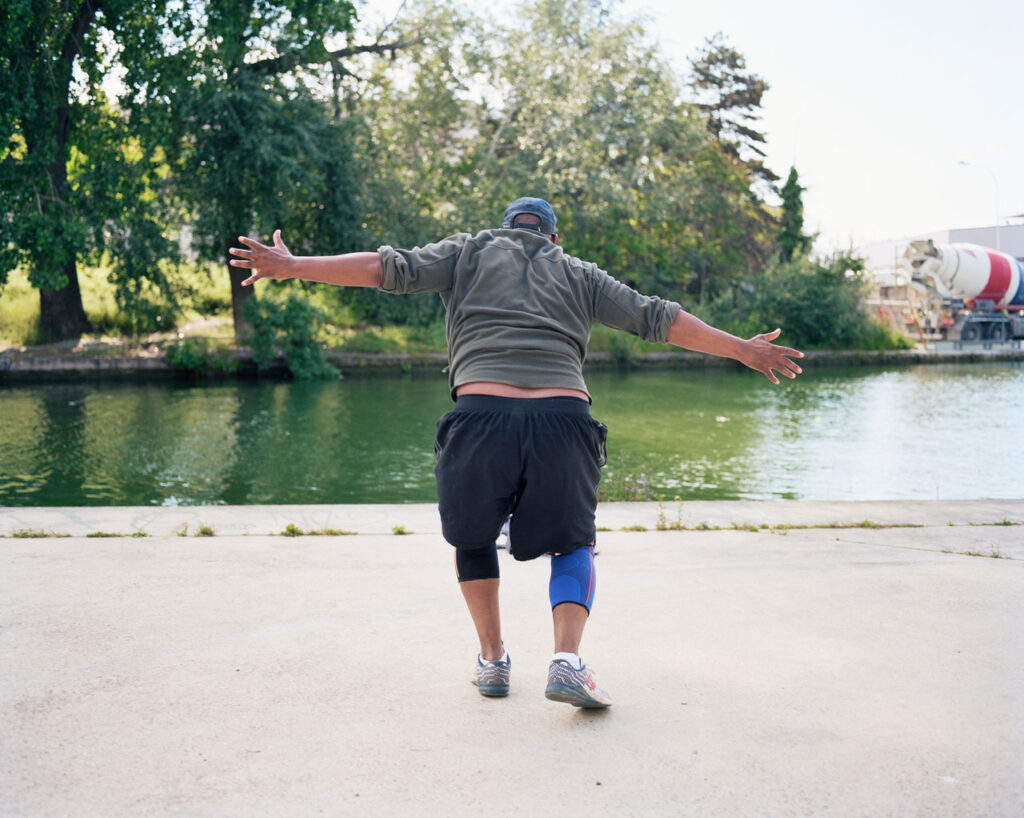 A man photographed from behind dances with open arms facing a canal in a Paris district.