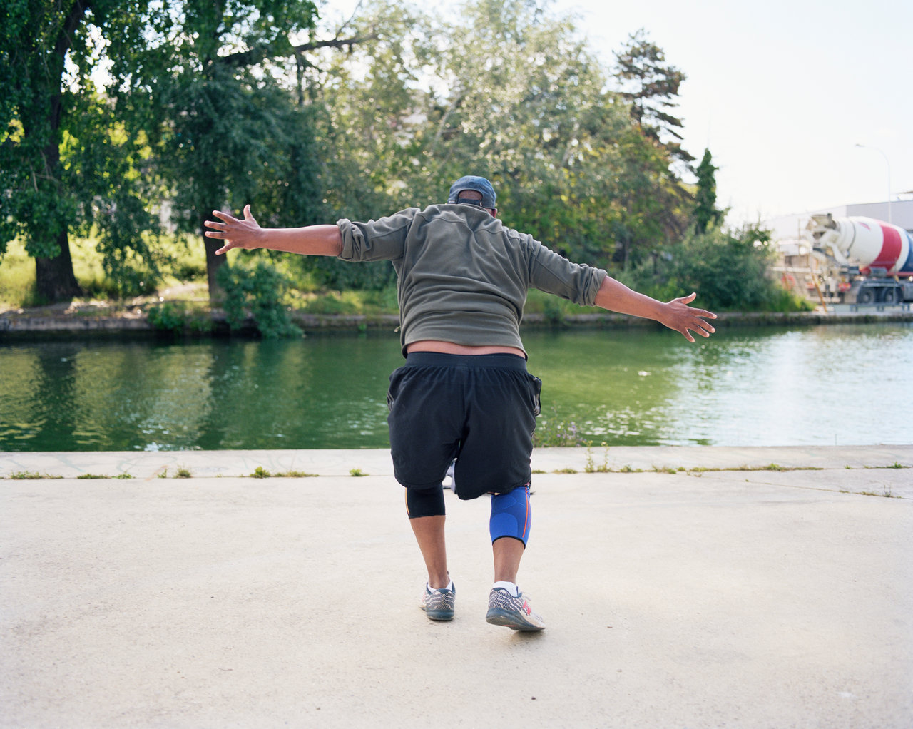 A man photographed from behind dances with open arms facing a canal in a Paris district
