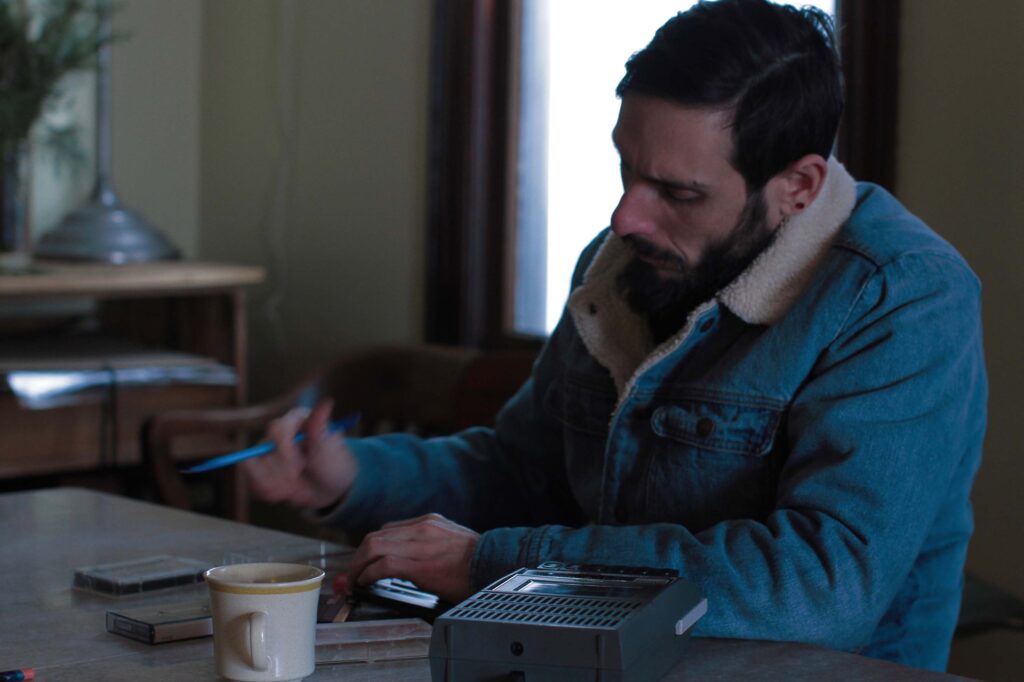 In a still image from a video, a man wearing a wool-lined jean jacket sits at a table or desk holding a pen with a coffee cup and cassette tapes scattered around him. He appears to be transcribing a tape recording to his left. 