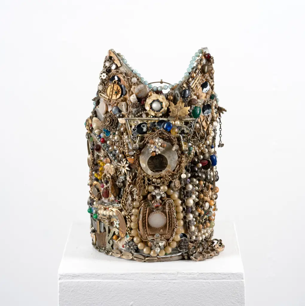 A crown shaped object on a pedestal. The crown is intricately encrusted with small gold charms, beads, pearls, and other small objects.