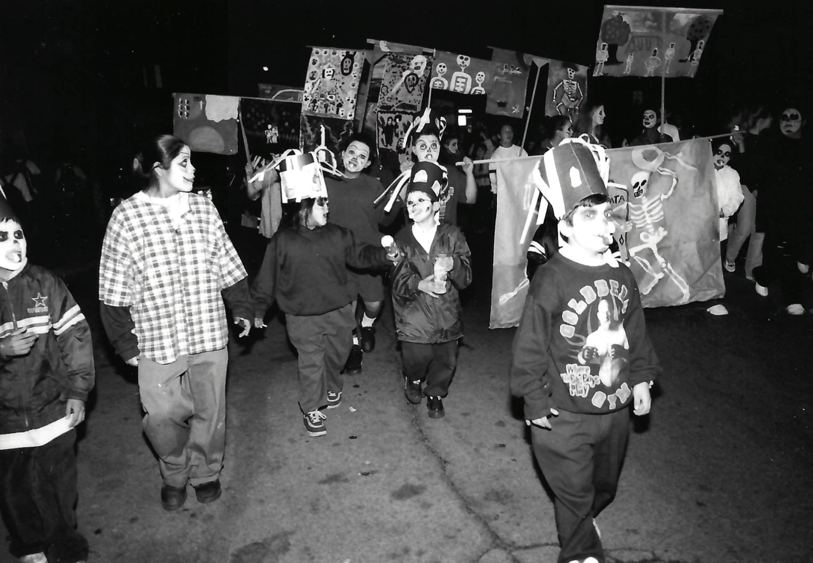 Children walking on a dark street wearing face paint and carrying signs and banners.