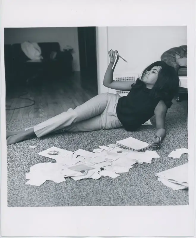 A woman with long hair reclines on carpet. A pile of letters are strewn about in front of her.