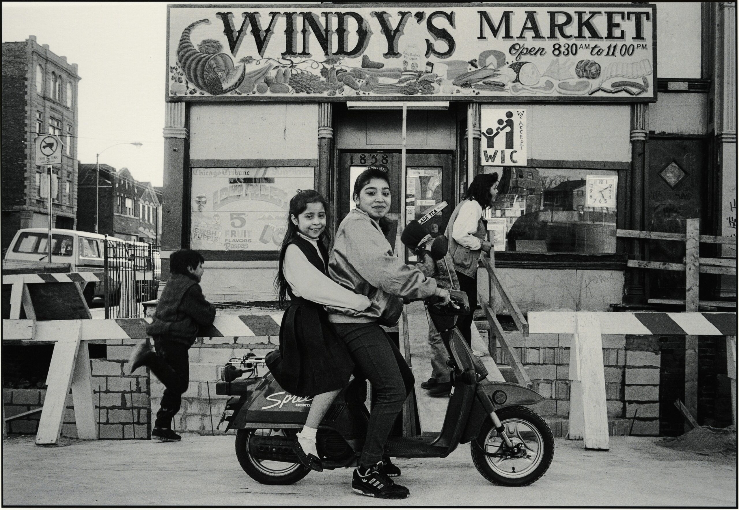Two young people seated on a scooter in front of a shop with a sign on top that says "Windy's Market."