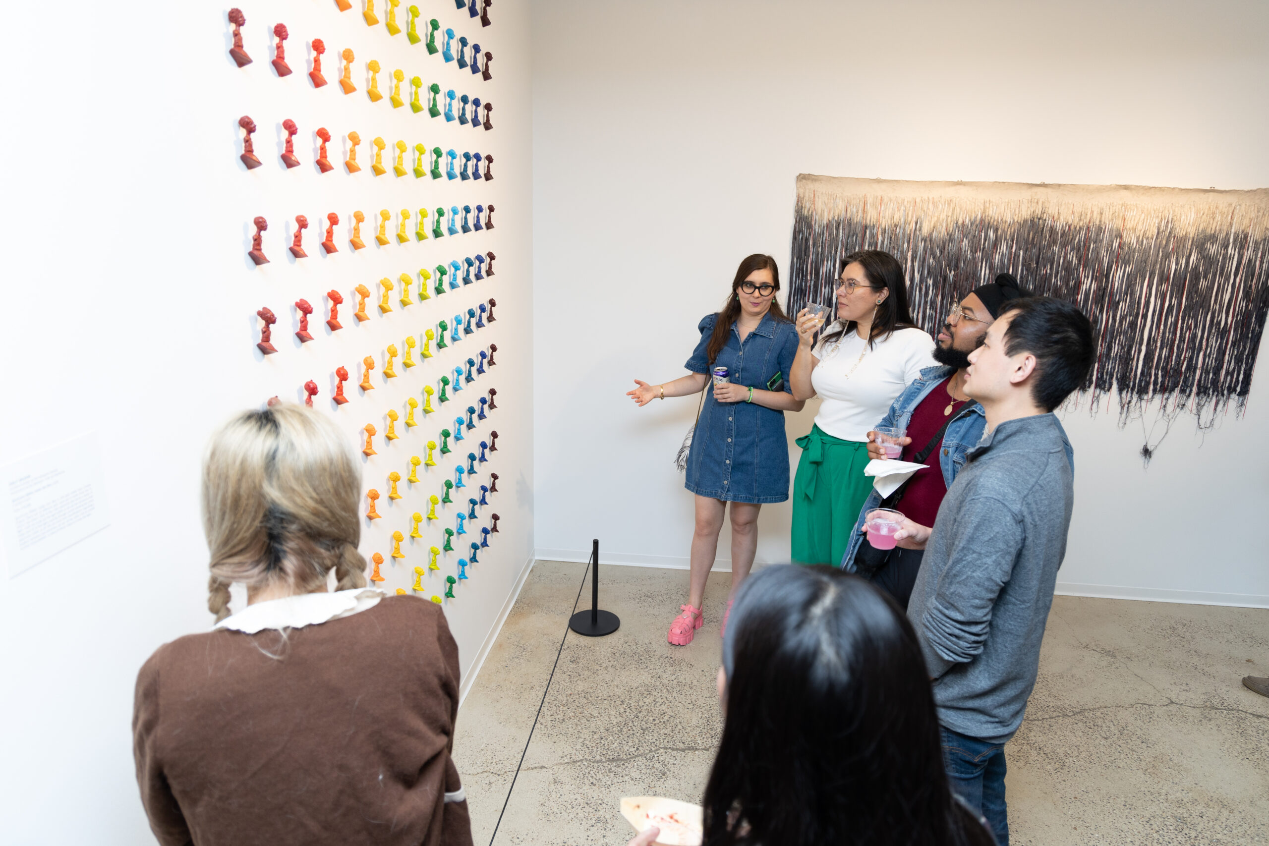 Six individuals stand in from of an artwork made up of small figurines in a rainbow of colors mounted on the gallery wall.