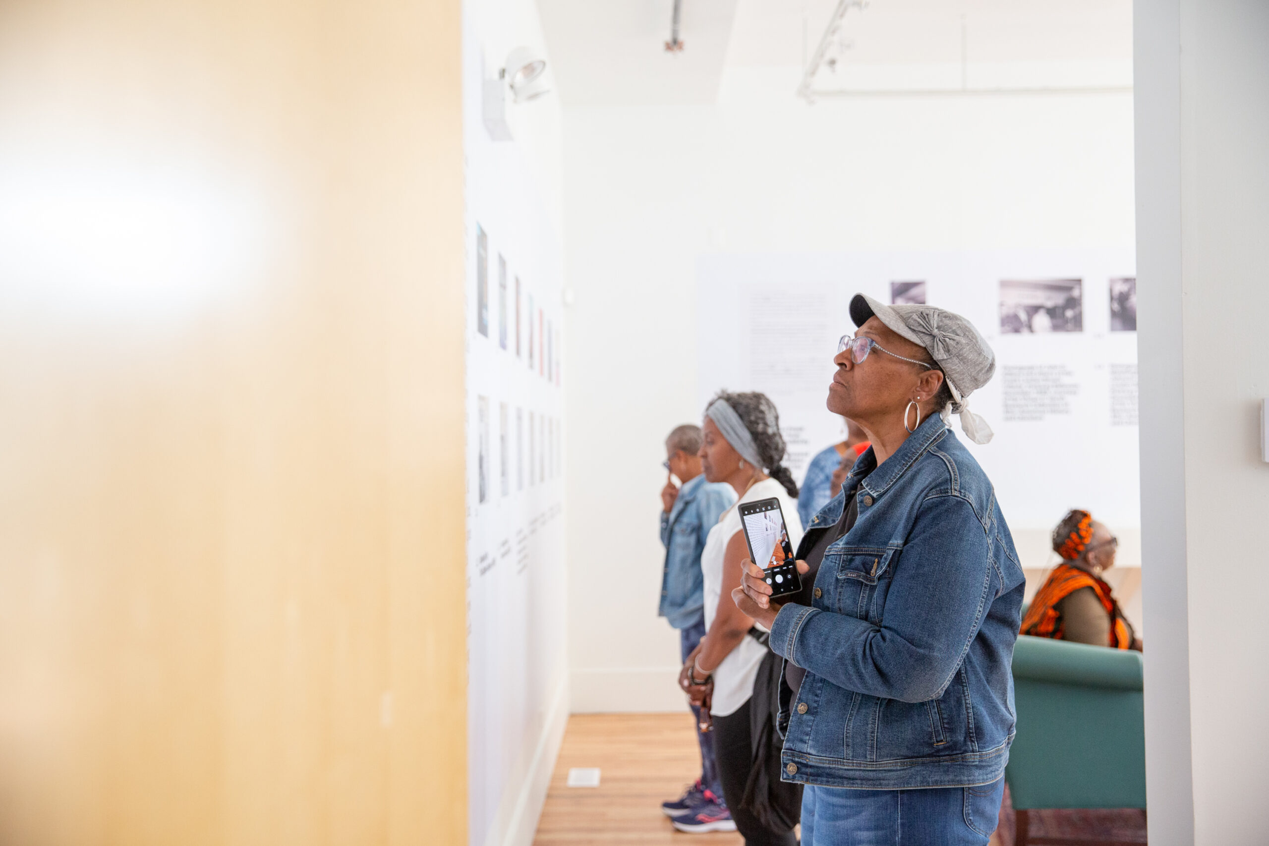 Visitors viewing artwork on a gallery wall. In foreground, visitor wearing a hat, hoop earrings, and a jean jacket. Other visitors in the background.