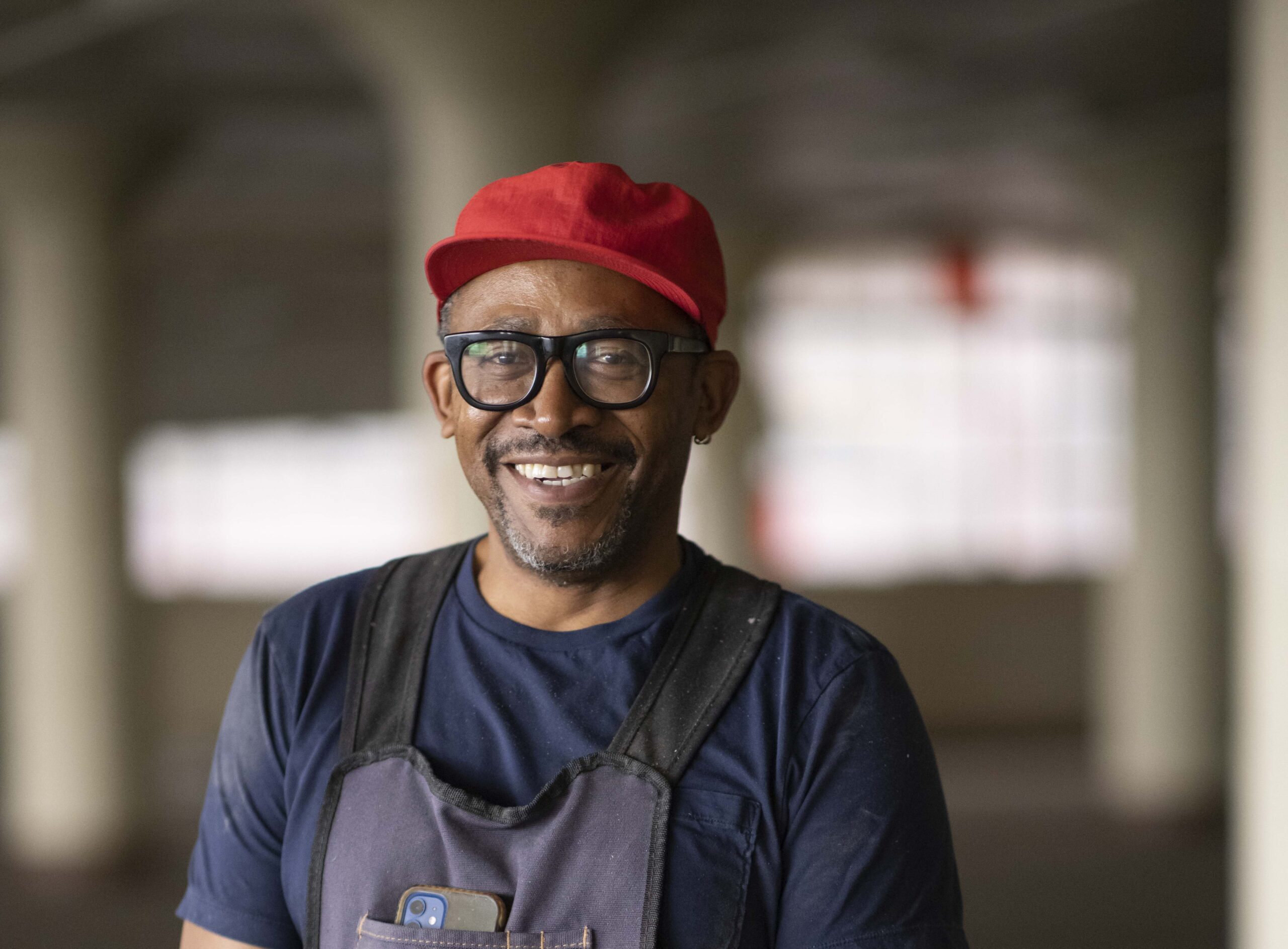 Portrait of Norman Teague wearing a red hat, glasses, blue t-shirt, and apron.