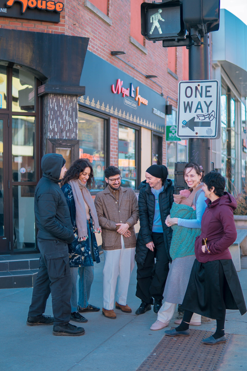 A group of six adults dressed in cold weather clothing stands on a Chicago street corner in front of a storefront and traffic signal pole.