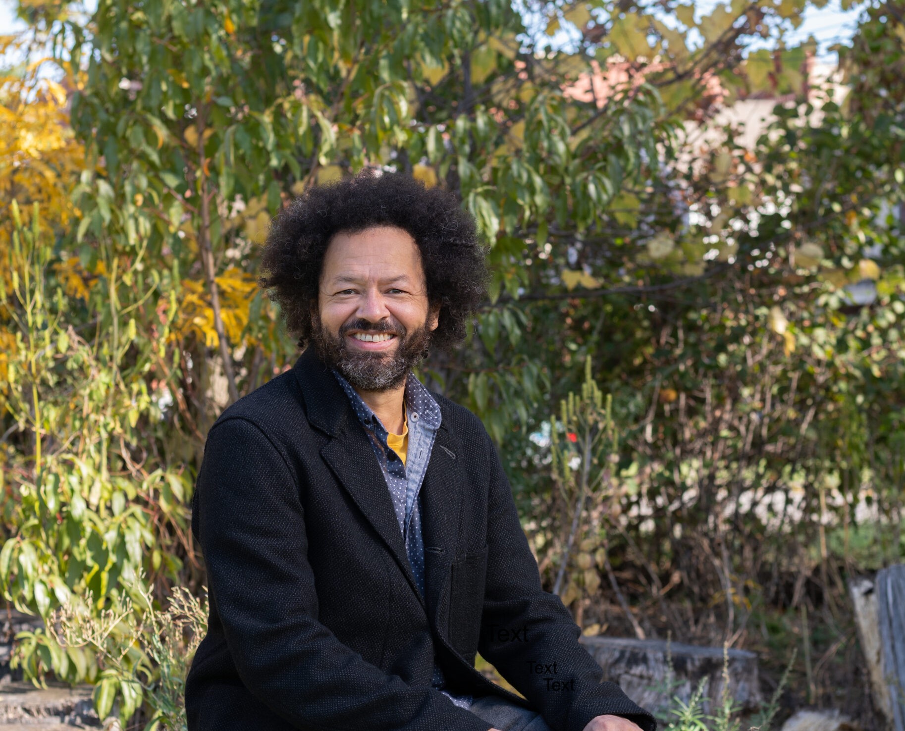 Portrait of David Brown seated in an outdoor space, smiling and wearing a black jacket, grey pants, and a blue shirt.