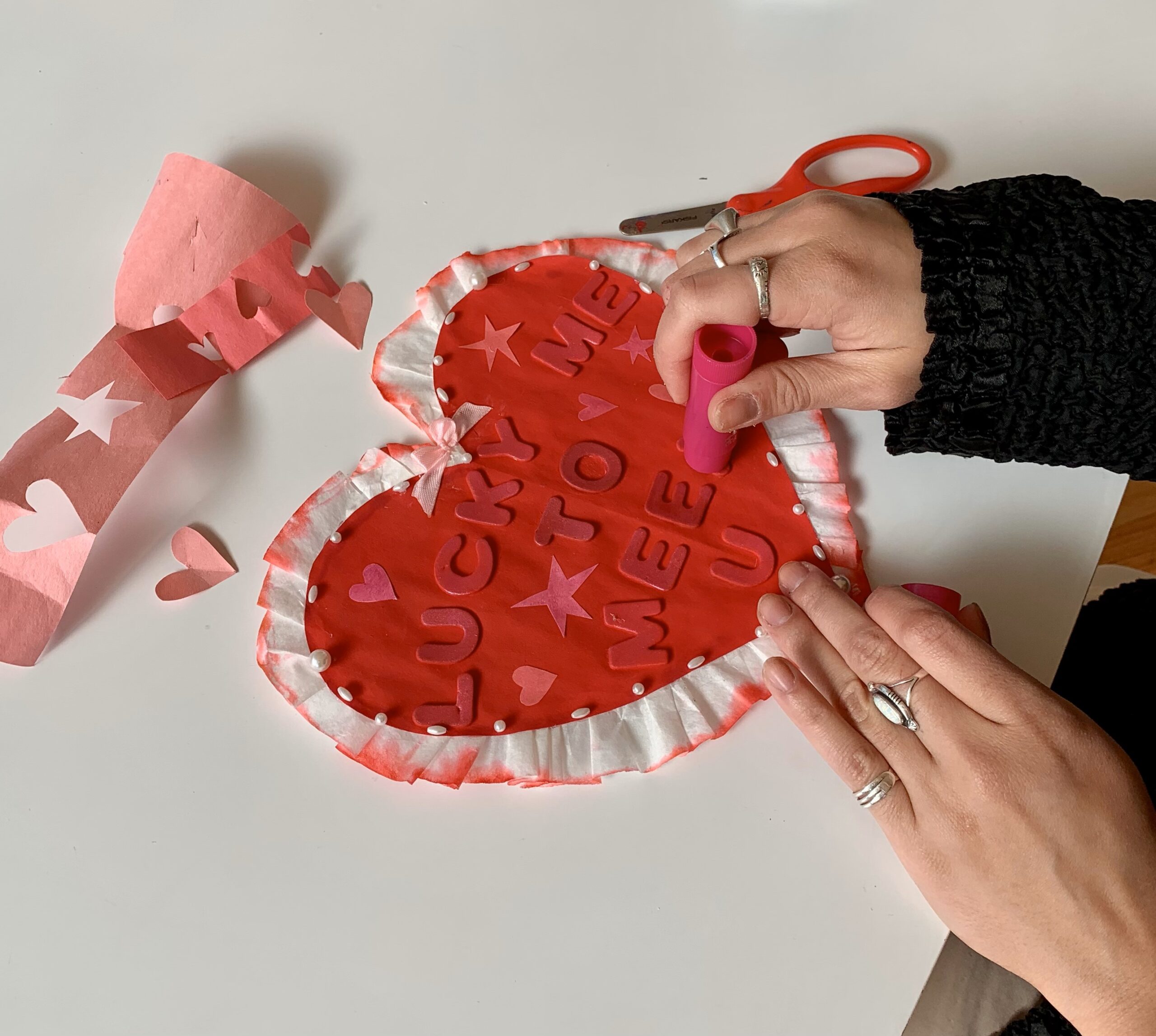 Hands of a person wearing rings and a black sweater working on a craft project. Project includes a paper heart with lettering that says "Lucky Me To Meet U."