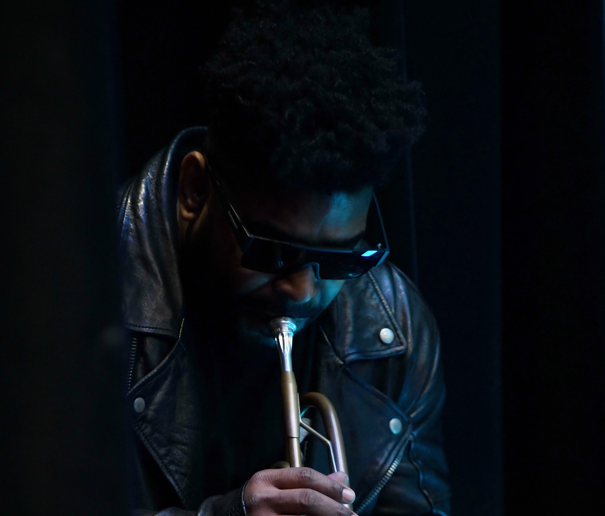 Portrait of Corey Wilkes wearing sunglasses, a black leather jacket, and playing a trumpet.