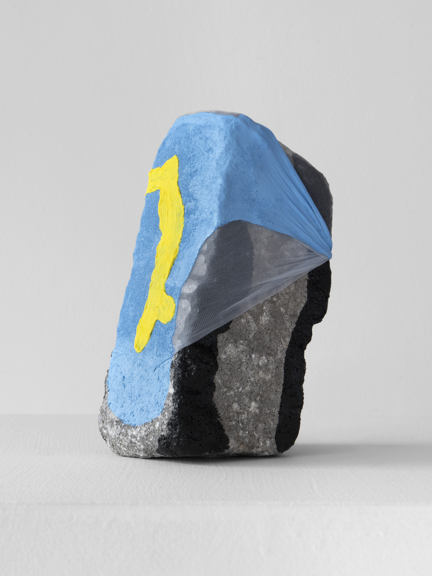 A sculpture in the form of a rock. Painted gray with black vertical stripes and covered in nylon painted blue and yellow.