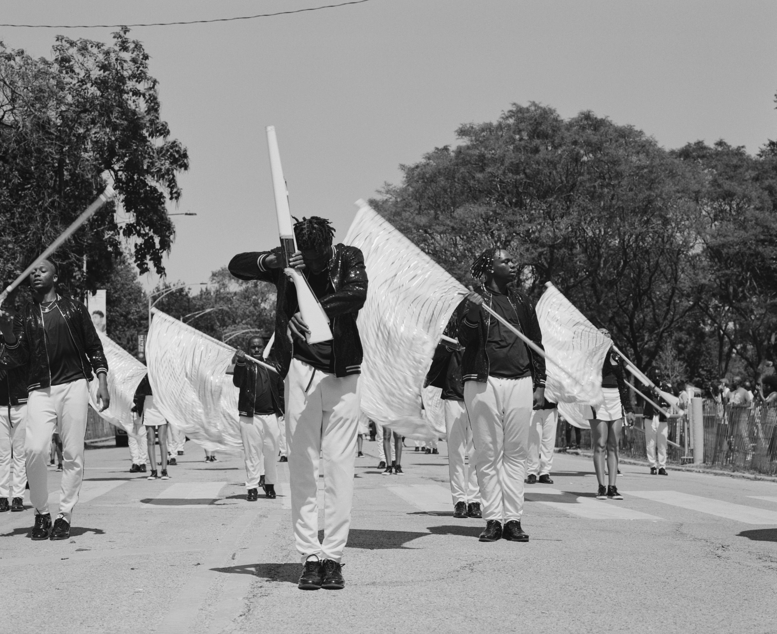 Photograph of South Shore Drill Team during a performance. Central figure is twirling a rifle while the figures in the back are performing with large white flags. 