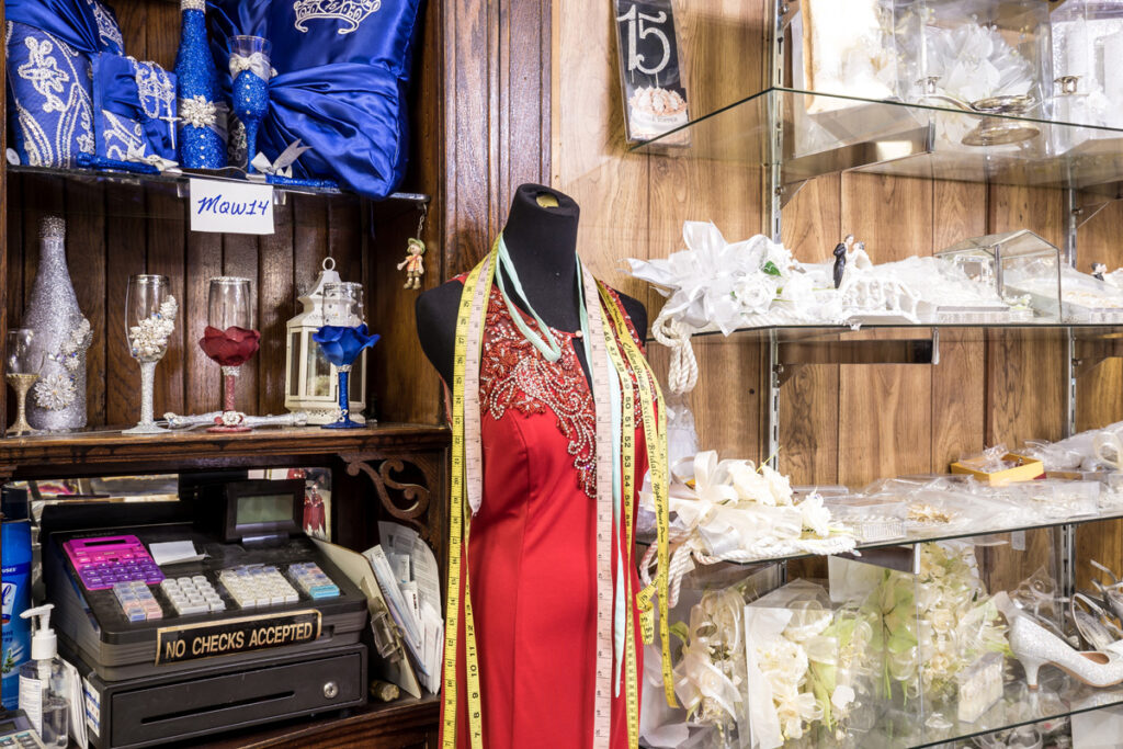 The interior of a shop features a headless mannequin wearing a red dress with beaded neckline and tape measures drapped around the neck. The the left and right are shelves containing glassware, a cash register, and many white wedding accessories.