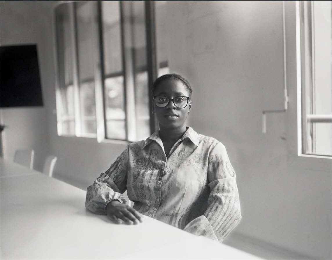 A young woman sits at a long table facing the camera. She wears a button down striped shirt and glasses.