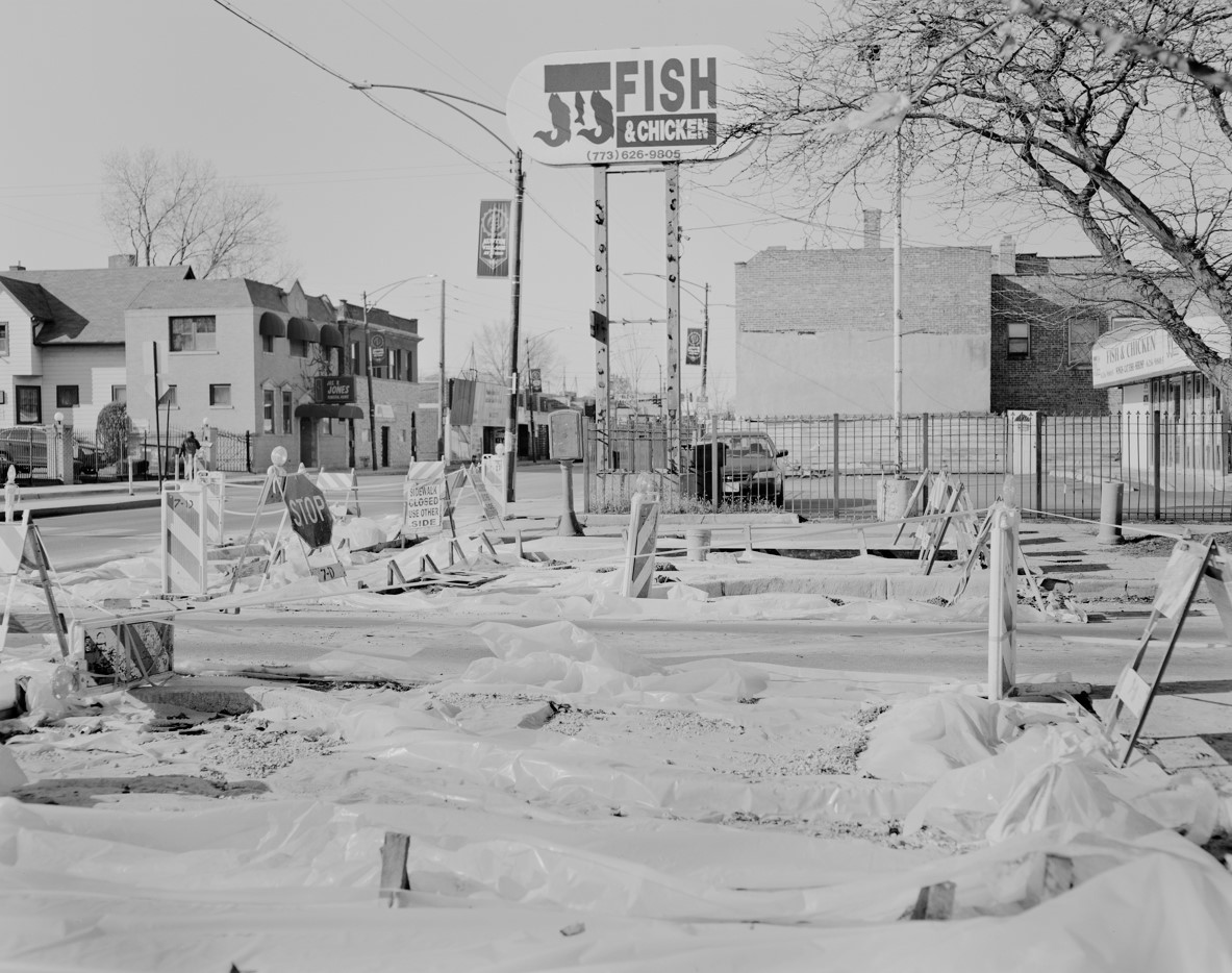 A black and white streetscape with an empty lot covered in plastic and traffic barriers in the foreground. In the background is a tall sign for JJ's Fish & Chicken.