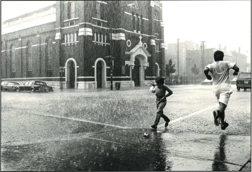 Two boys run through a very rainy Pilsen street corner. A large church and cars in the background.