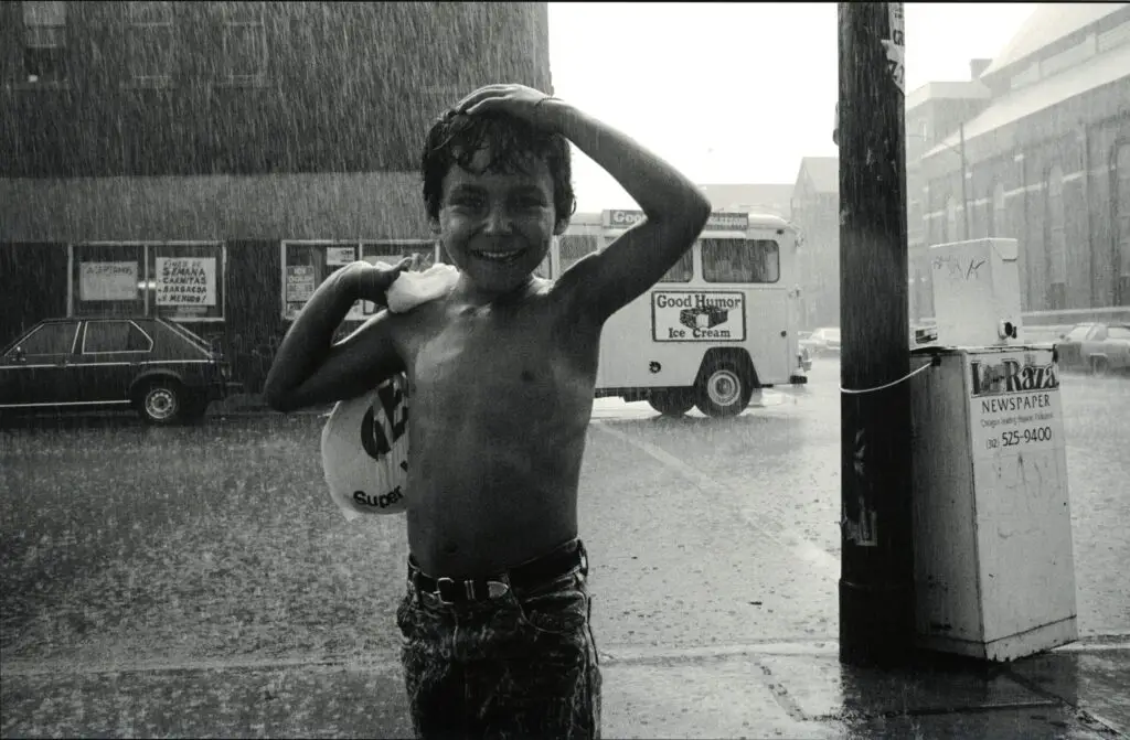 A young boy, shirtless, stands in the rain, hand on head, with a big smile. An ice cream truck is in the background.