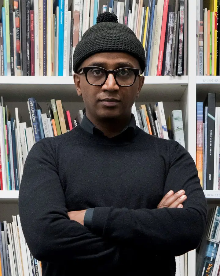 Dawit L. Petros stands in front of a bookshelf, arms crossed, wearing glasses and a black hat, shirt, and sweater.