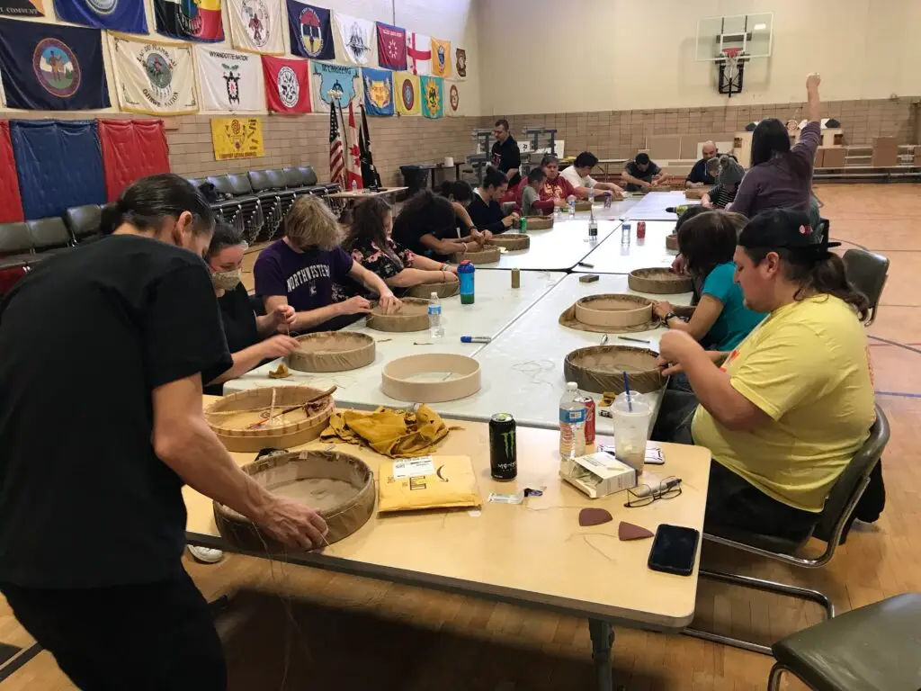 A large group of workshop participants sits at long tables in a gymnasium working on their hand drum projects.