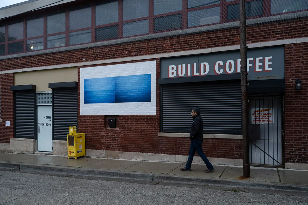 A street scene featuring an artwork pasted up on a brick wall. A figure passes by. A sign on the wall says "Build Coffee."