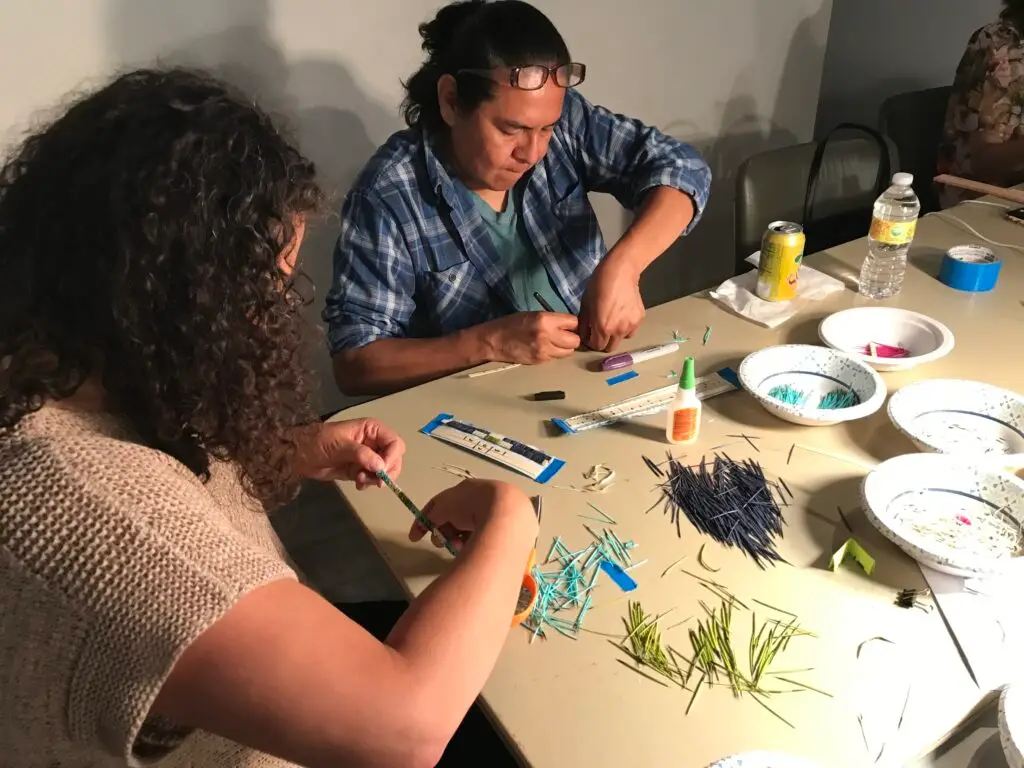 Two workshop participants are working at a table covered with colored quills and other materials.