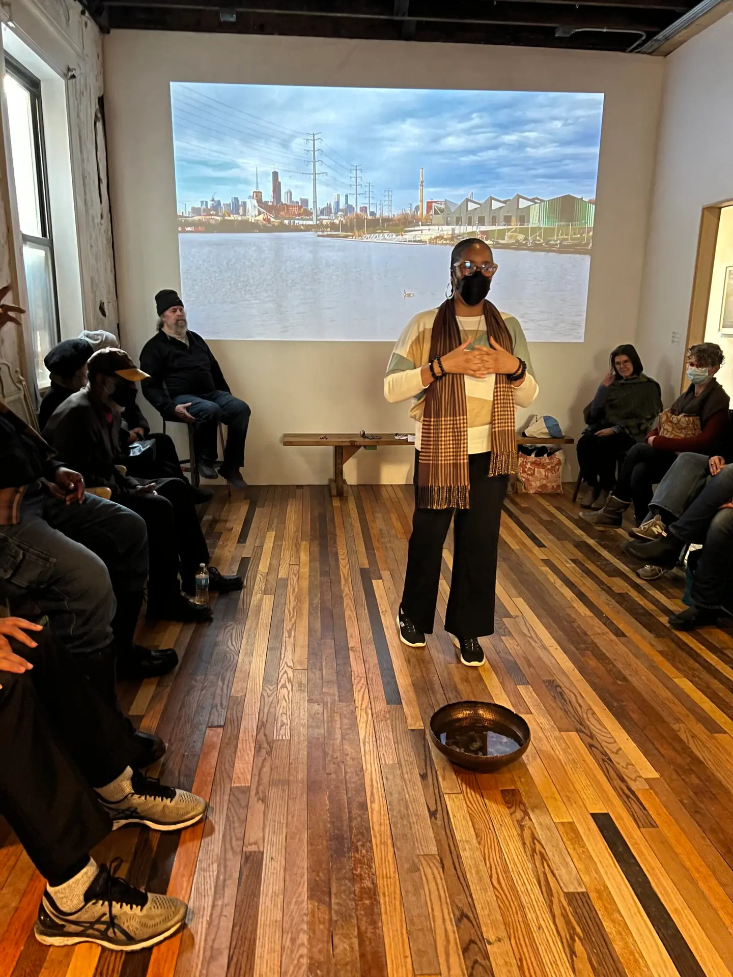 A workshop facilitator stands at the center of a room with a metal bowl filled with water at her feet. Participants are seated against the walls. 