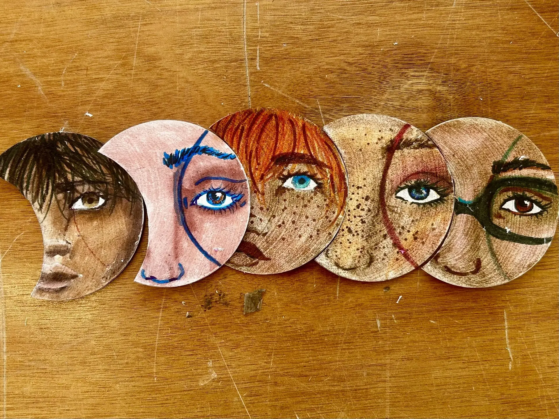A series of five overlapping crescent-shaped paintings of faces overlap on a wood tabletop.