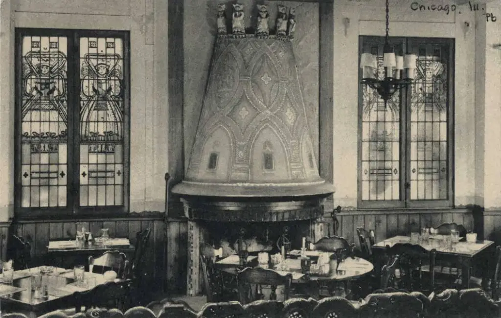 Black and white photograph of a large room with decorative fireplace, restaurant tables, and large stained glass windows