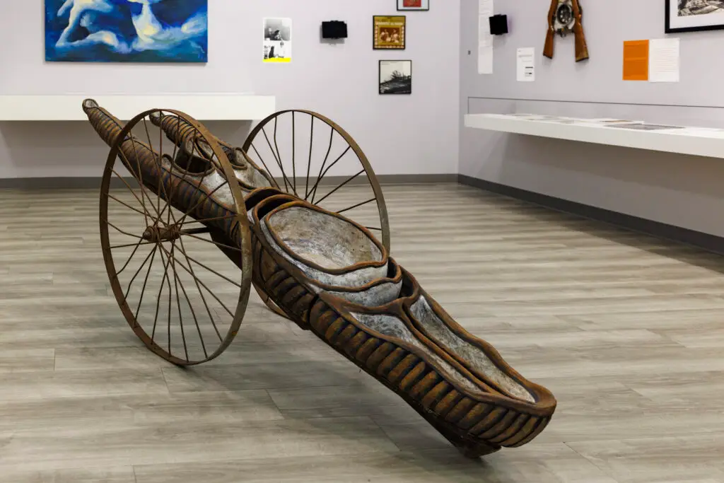 An abstract sculpture with a long central object and two large thin wheels sits on a gallery floor.