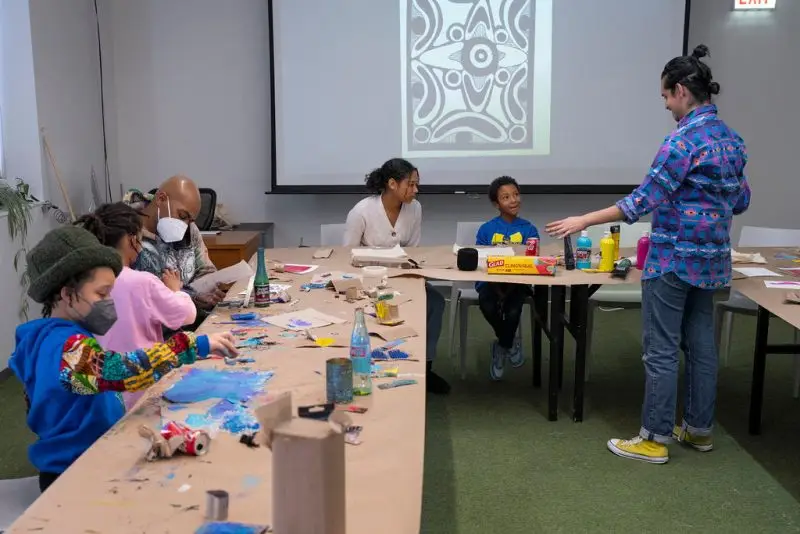 A group sits at a table covered with brown paper and craft supplies working on a project. The instructor stands in front of them.