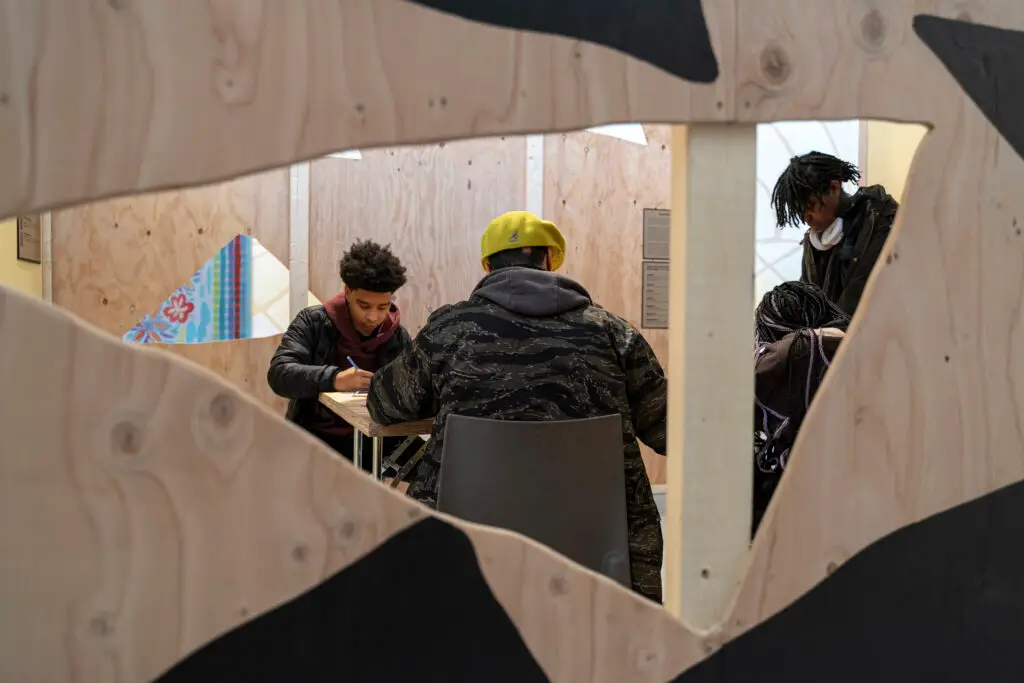 View looking inside a wooden structure at a group of young people seated at a table working on a craft project.