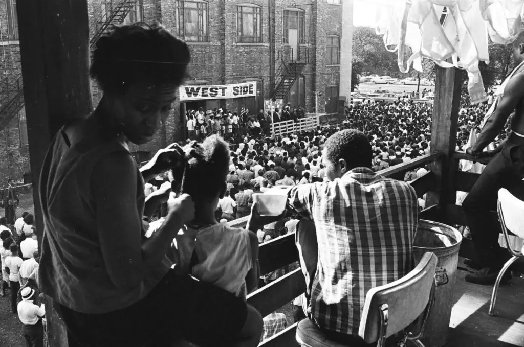 A family sits on a balcony above a very crowded street filled with people. A banner across the street reads "West Side."