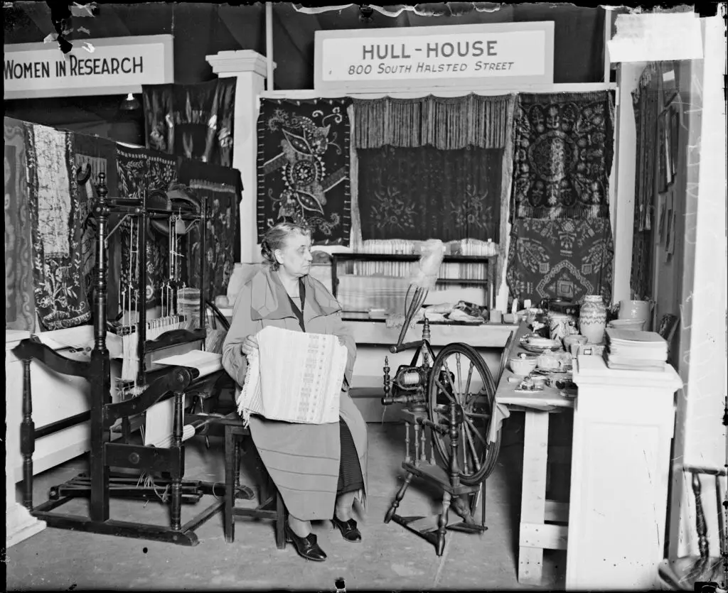 Black and white photo of an older woman (Jane Addams) seated amongst woven and sewn textiles, a spinning wheel, and a weaving loom. There is ceramic pottery on a table nearby. 