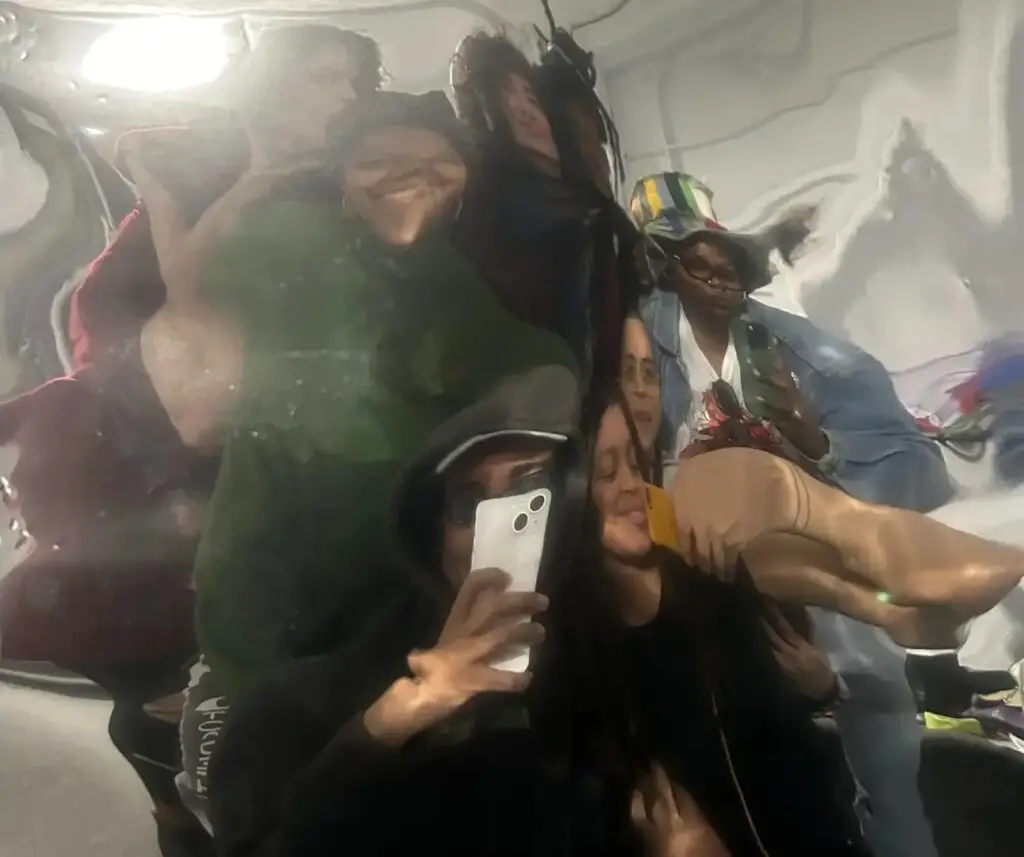 A group photo in a funhouse mirror, distorting a group of seven artists in a white exhibition space, with an iPhone held up in the center taking the photograph.