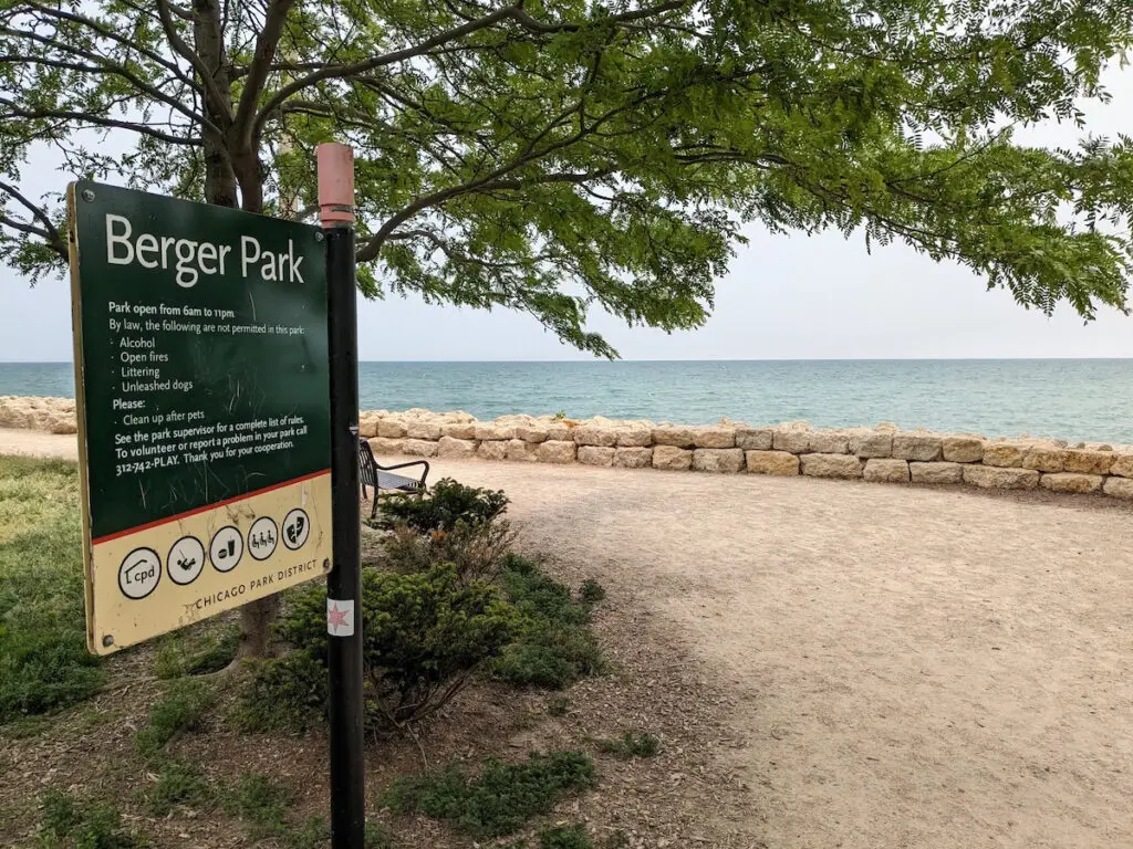 A green and tan park district sign with a list of rules is posed at an angle to the blue water of Lake Michigan.