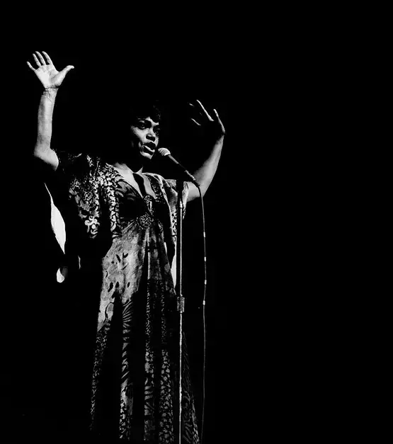 Portrait of Earth Kitt standing in front of a microphone, wearing a long metallic dress, hands in the air.