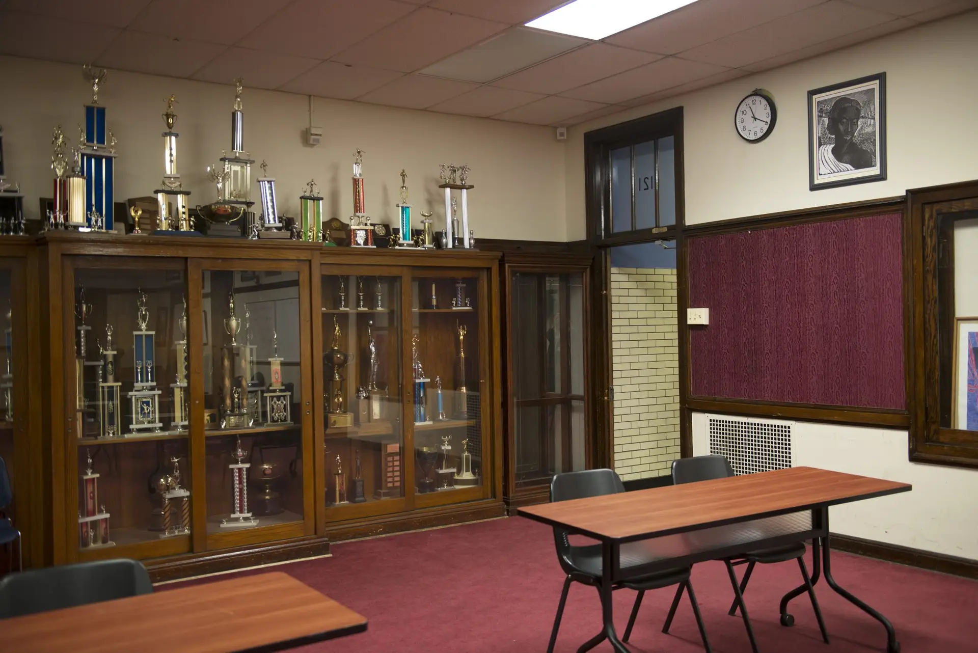 Photo of a classroom, empty of students, containing a large trophy case, tables, and chairs.