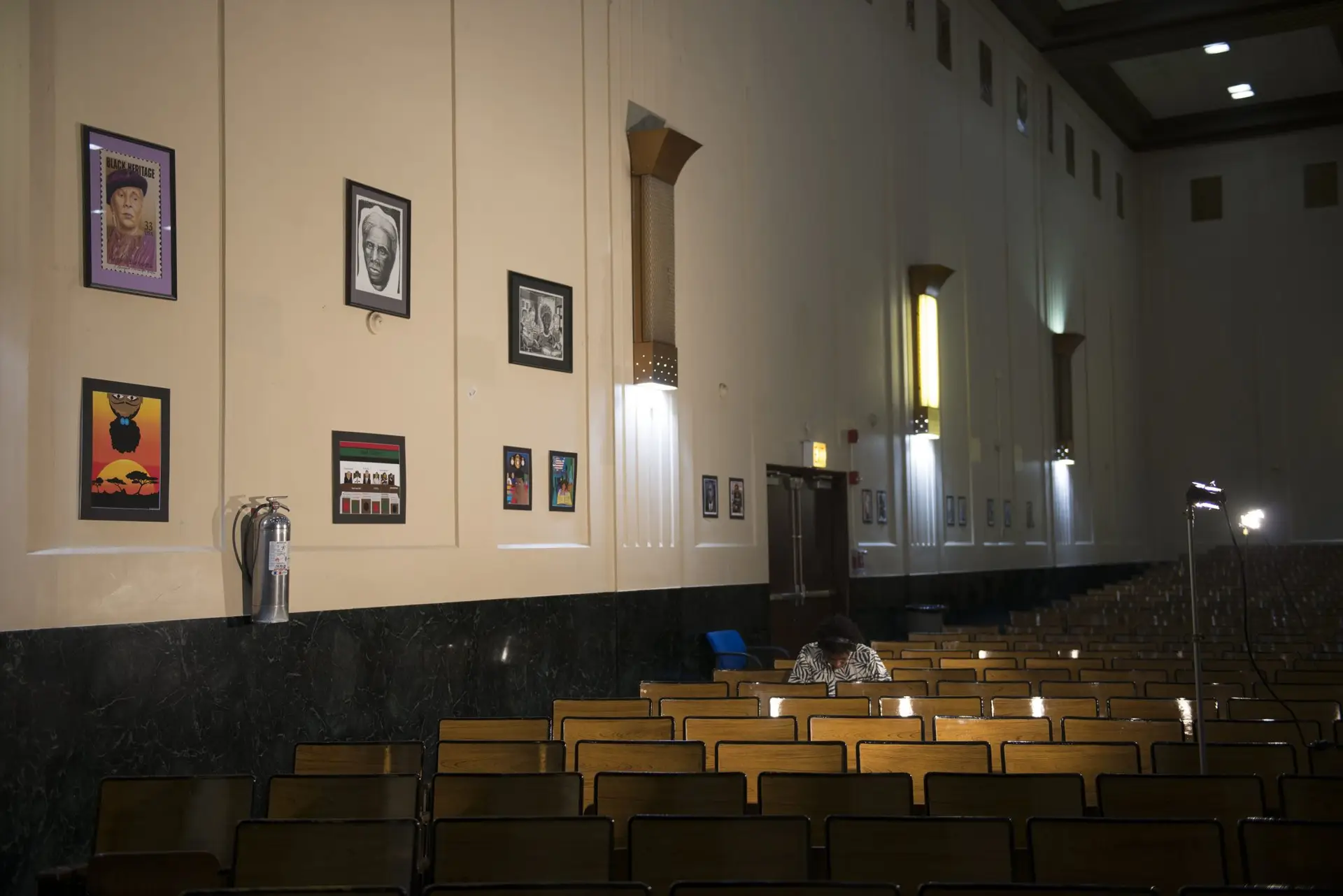 A student sits in a darkened school auditorium. Artwork and a fire extinguisher are on the wall.