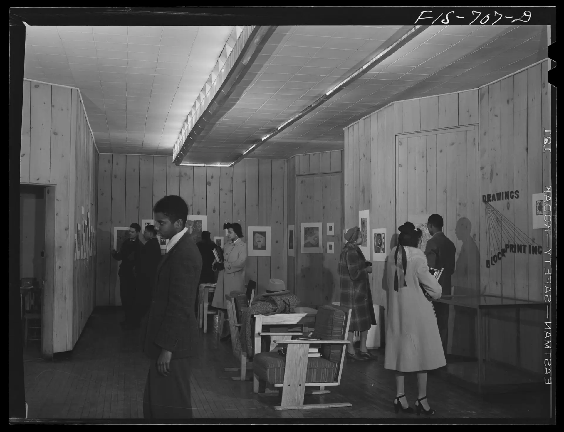 A vintage photo of people inside a small gallery space looking at art hung on wood paneled walls. Large chairs are positioned in the center of the gallery.