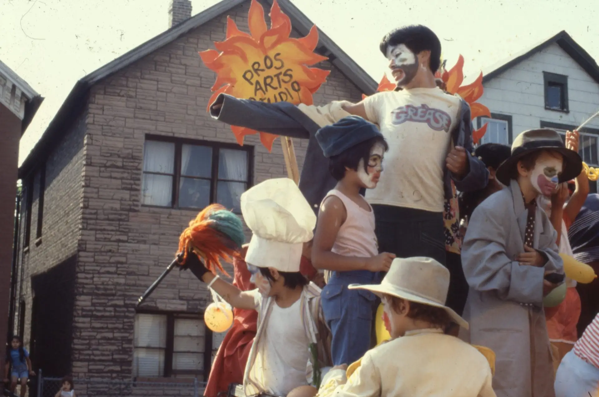 A group of children wearing costumes and face paint crowd together in front of a sun-shaped sign that says "Pros Arts Studio."