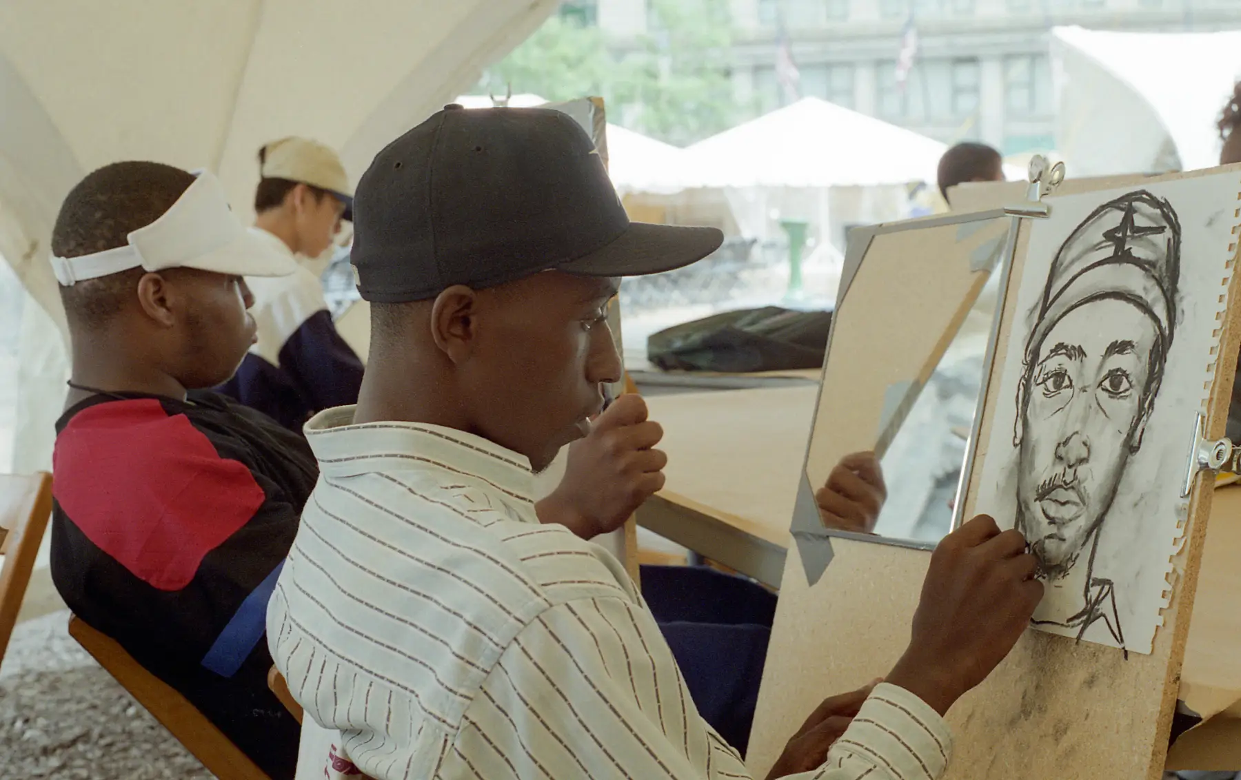 A person wearing a hat and striped shirt sits drawing a portrait. Other artists are seated in the background.