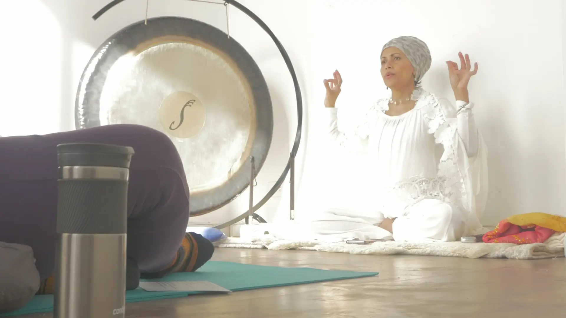 Aneela Dean is seated on the floor with legs crossed next to a large gong. She wears all white, with a beaded necklade and headcoving.